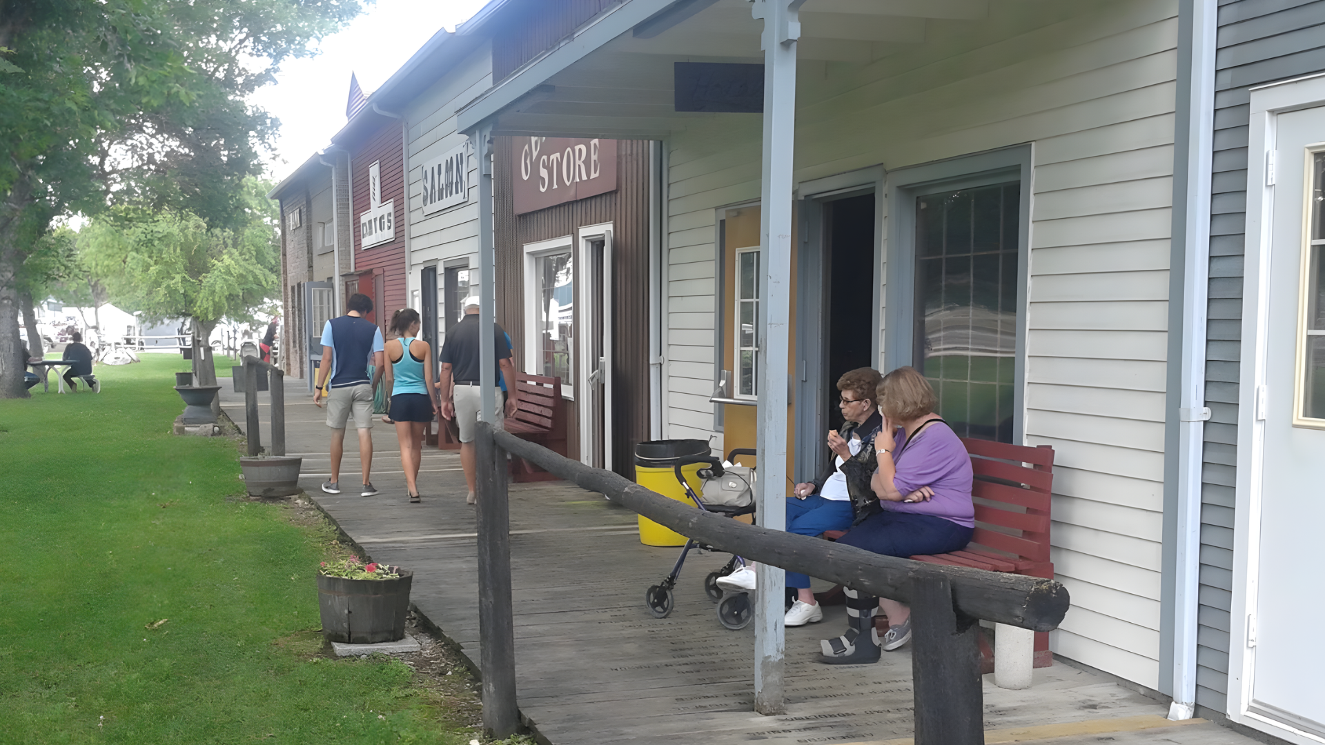 A row of storefronts with a wooden porch where people walk and sit on a bench on a sunny day.
