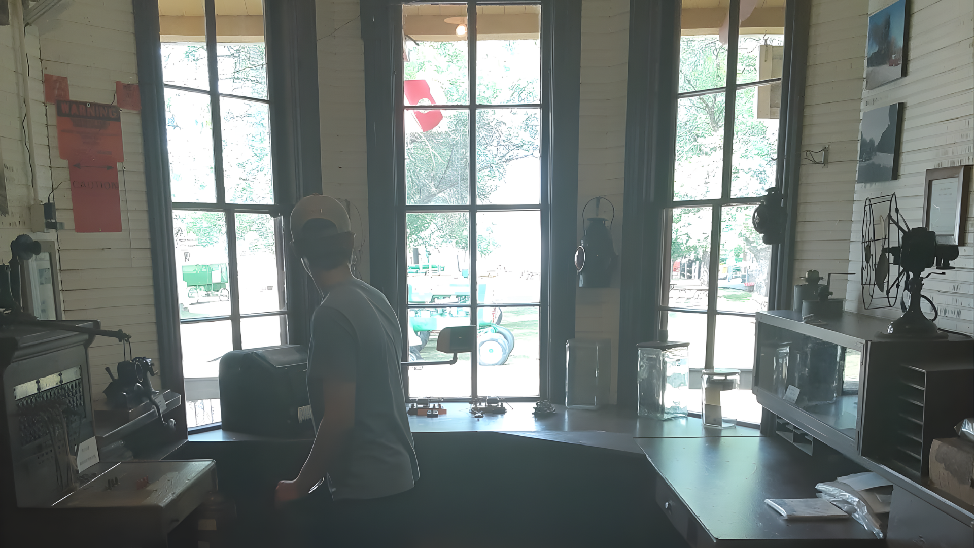 A person stands at a desk in a room with three large, bright windows overlooking an outdoor area.