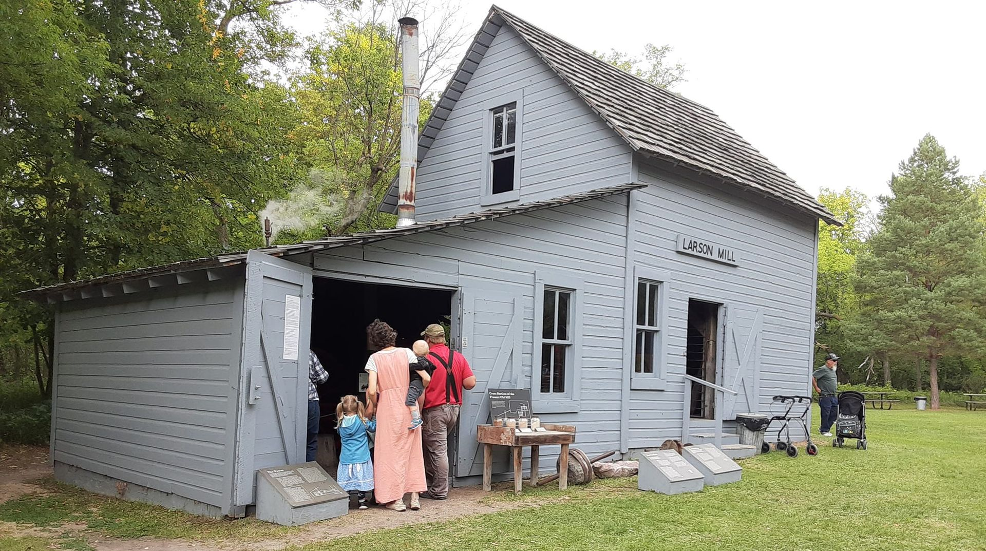 Visitors observe an old blue wooden house with a shingled roof, chimney, and an attached side structure in a grassy field.