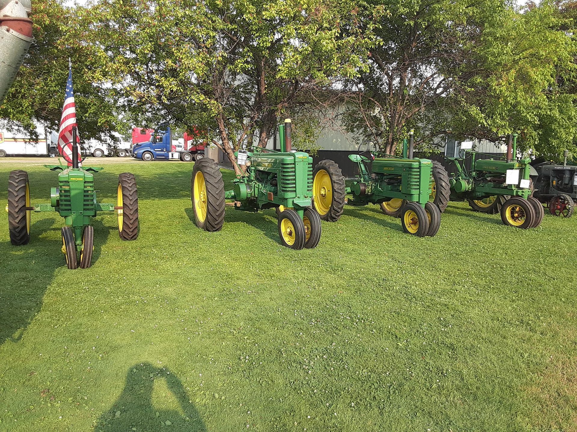 Four green and yellow John Deere tractors lined up on a grassy field, American flag in the background.