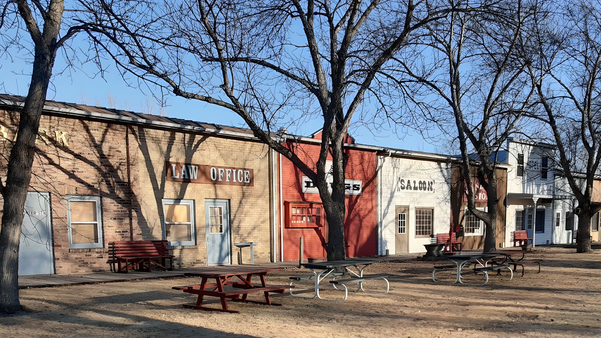 A row of old-west style storefronts with wooden picnic tables in a sunny, tree-lined courtyard.