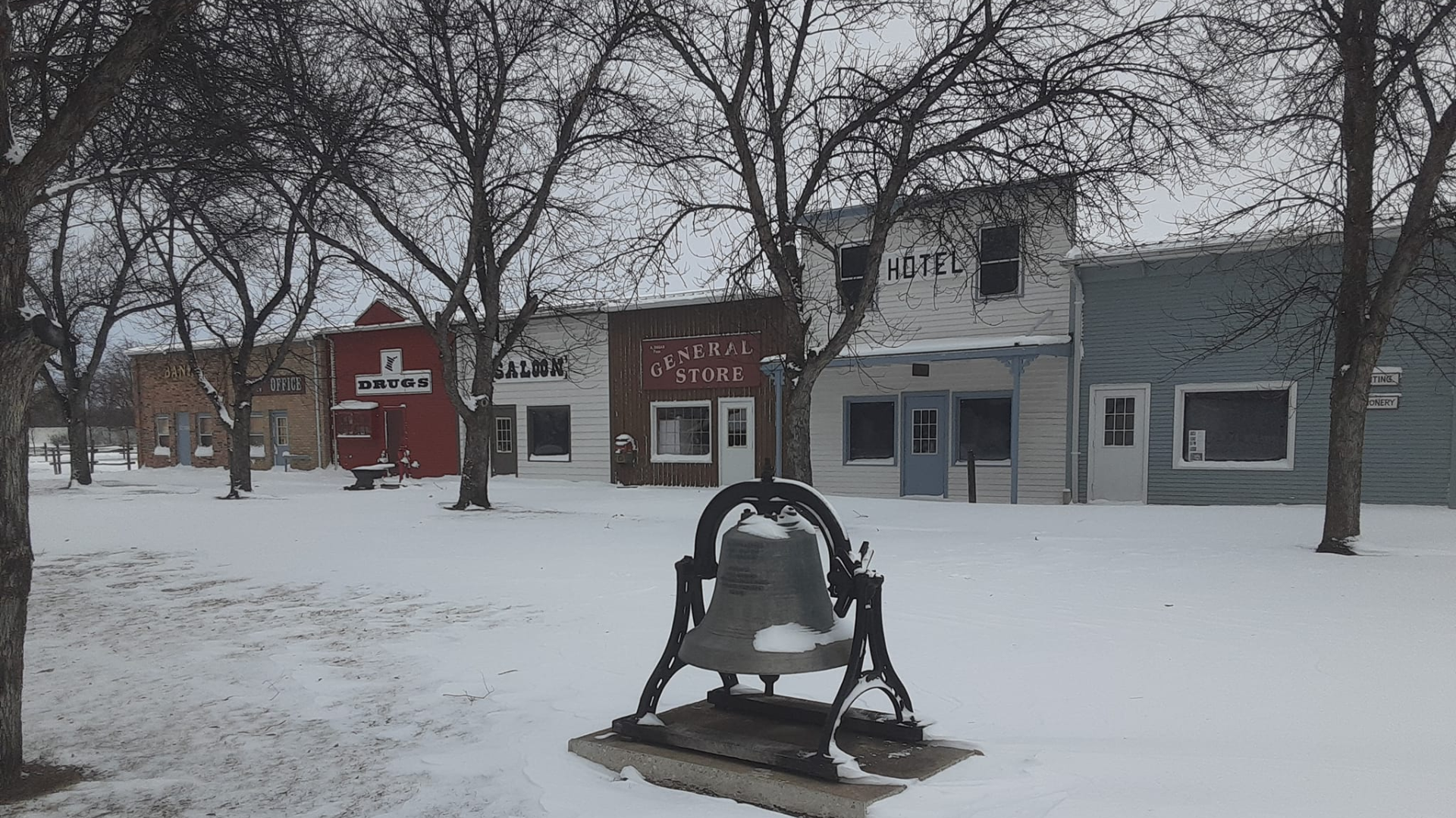 A snow-covered town square featuring a prominent, centrally placed iron bell, with a row of historic buildings behind.