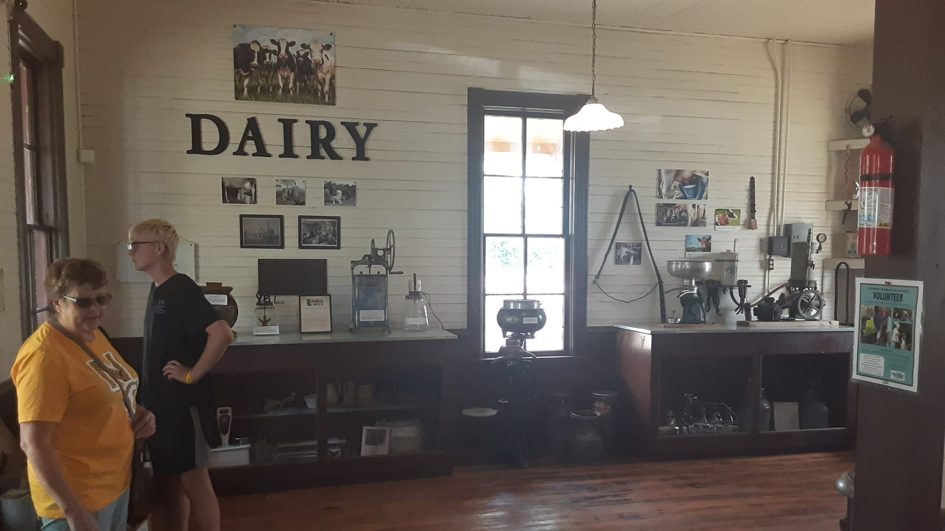 Two people stand in an old dairy display room with wood walls, vintage equipment, photographs, and a fire extinguisher.