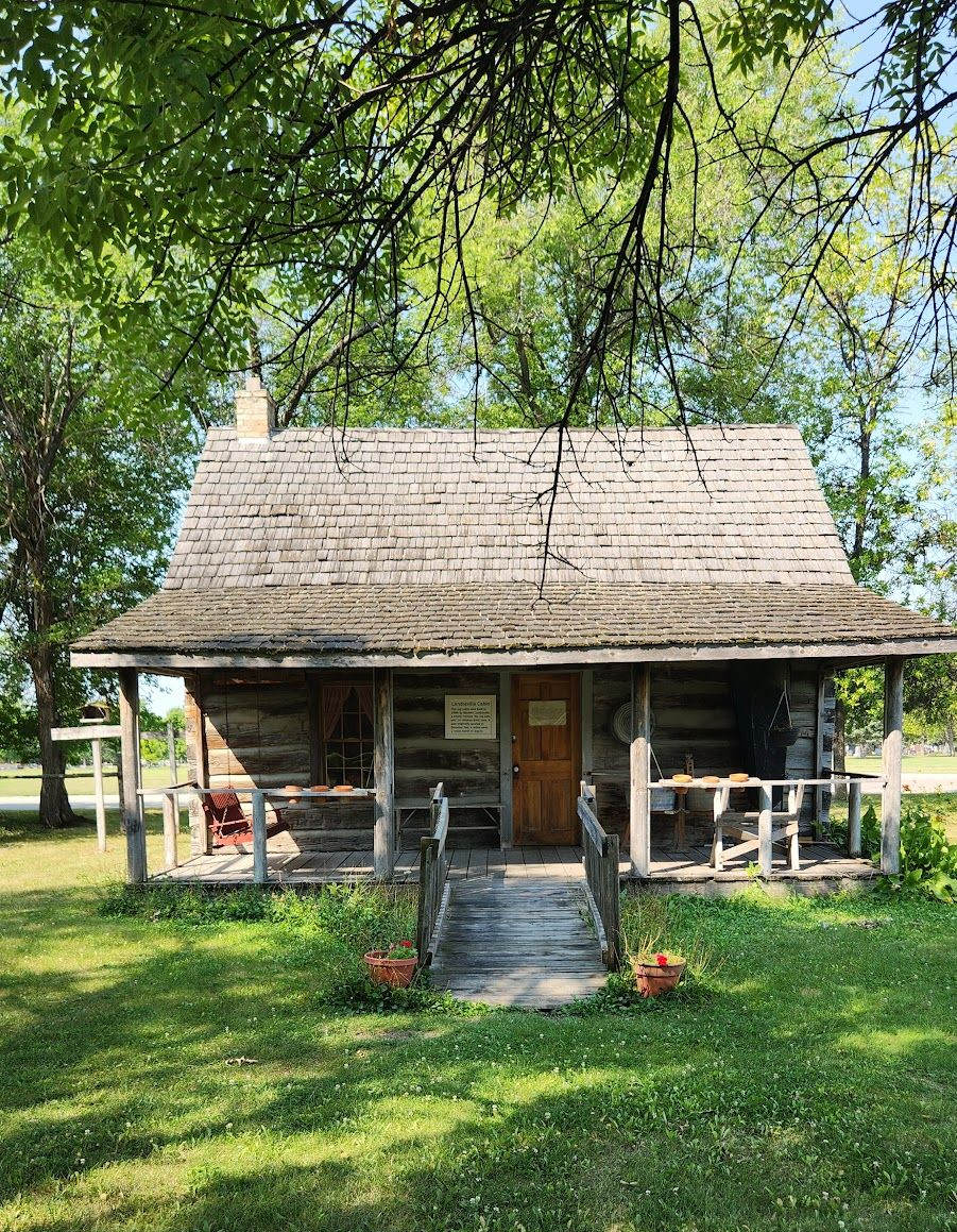 A rustic log cabin with a wooden porch and cedar shake roof, surrounded by green trees and a grassy lawn.
