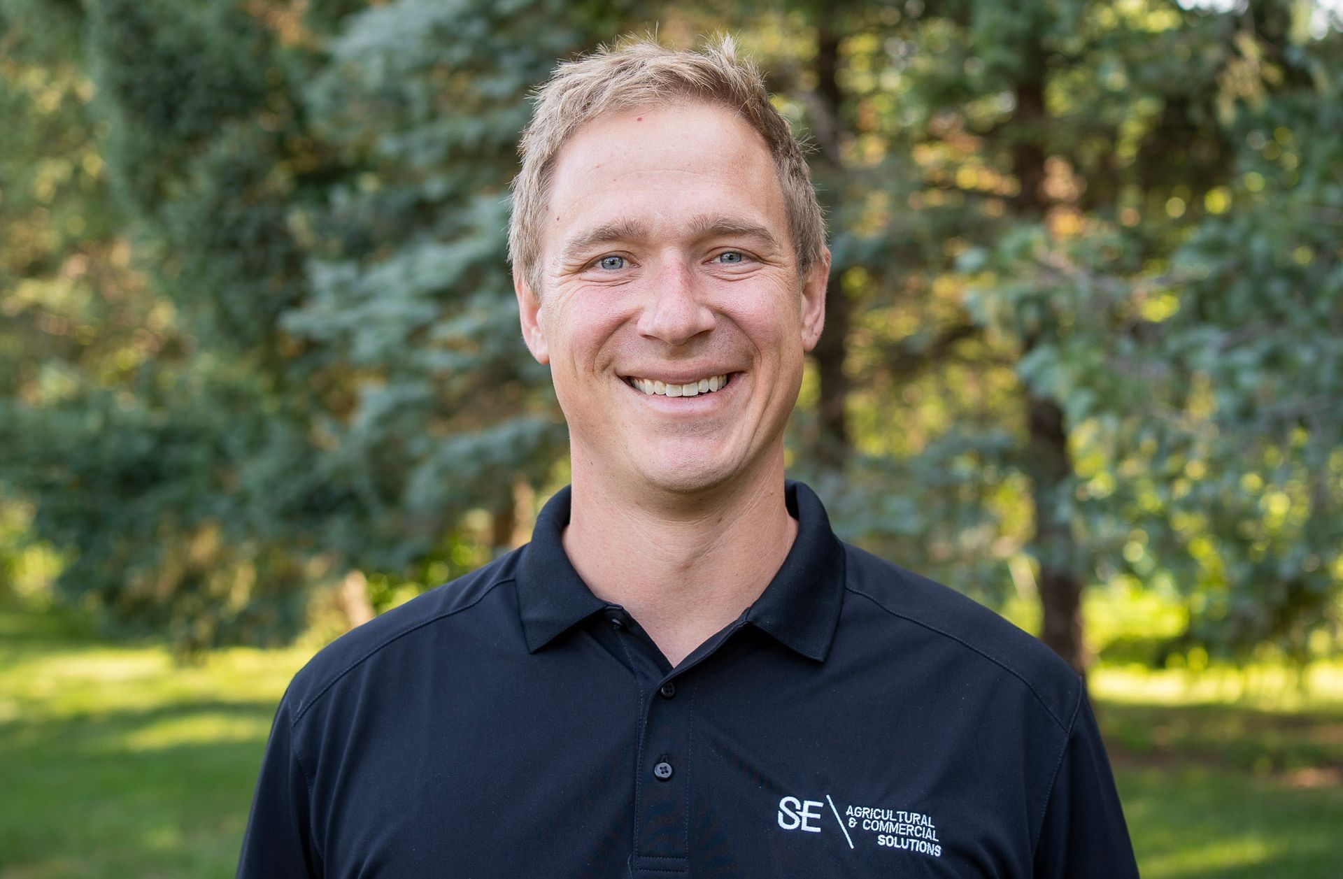 A smiling person in a black polo shirt stands in front of a soft-focus background of green trees and foliage.