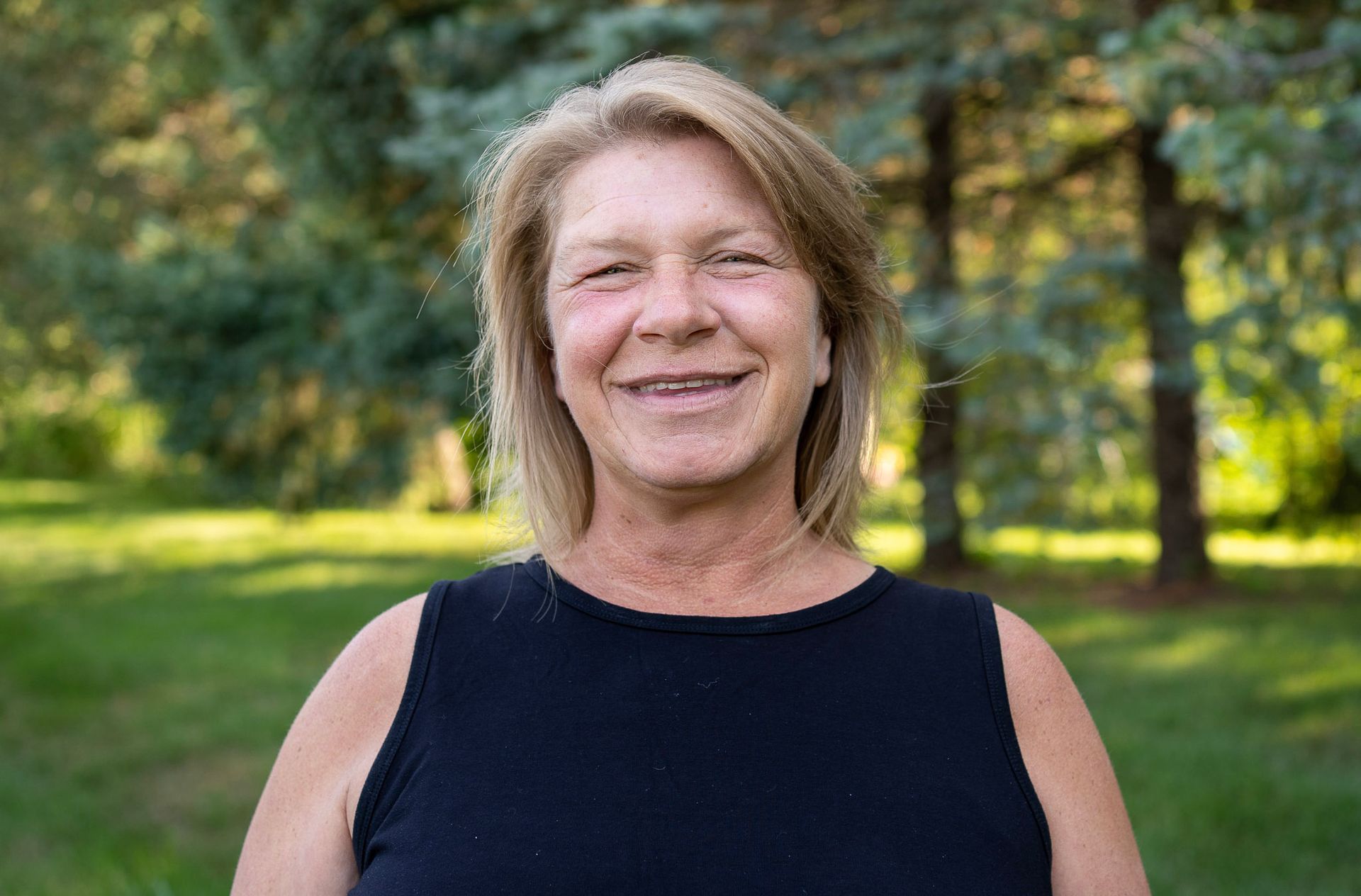 Smiling person with blonde hair wearing a black sleeveless top, standing outdoors in front of green trees.