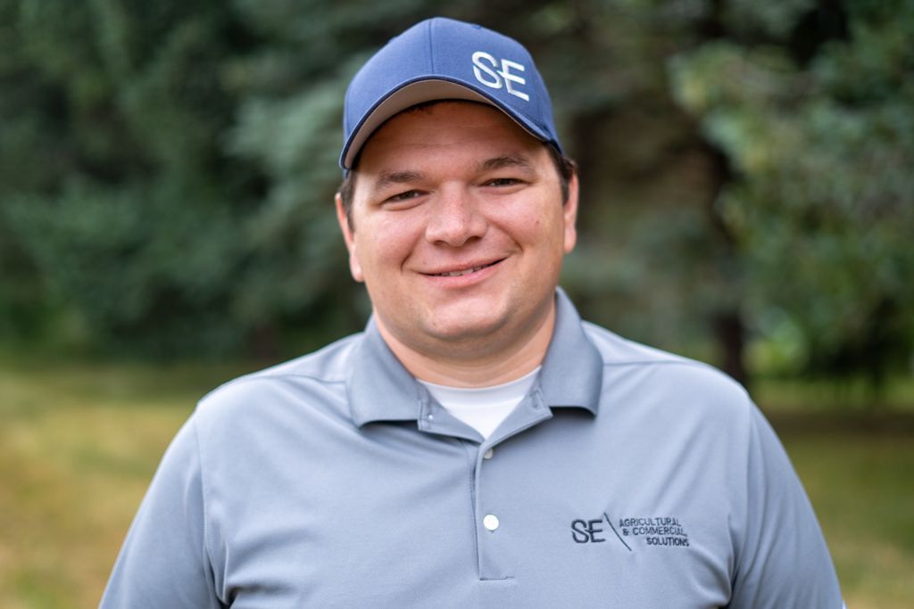 A person smiling, wearing a gray polo shirt and a navy cap with the letters