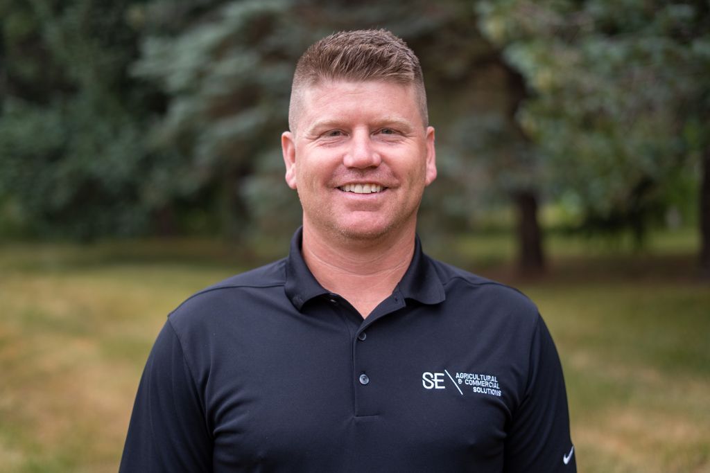 A person smiling while wearing a black polo shirt with a logo, standing outdoors in front of a blurry, green, wooded background.