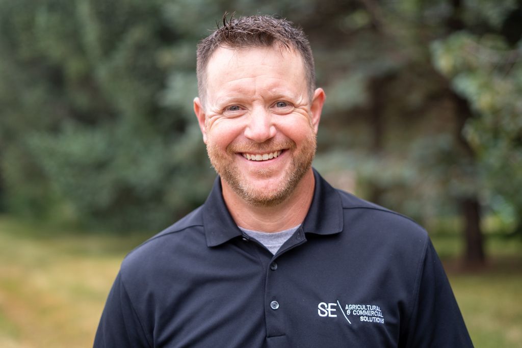 A person wearing a black polo shirt with a company logo smiles while standing outdoors against a blurred forest background.