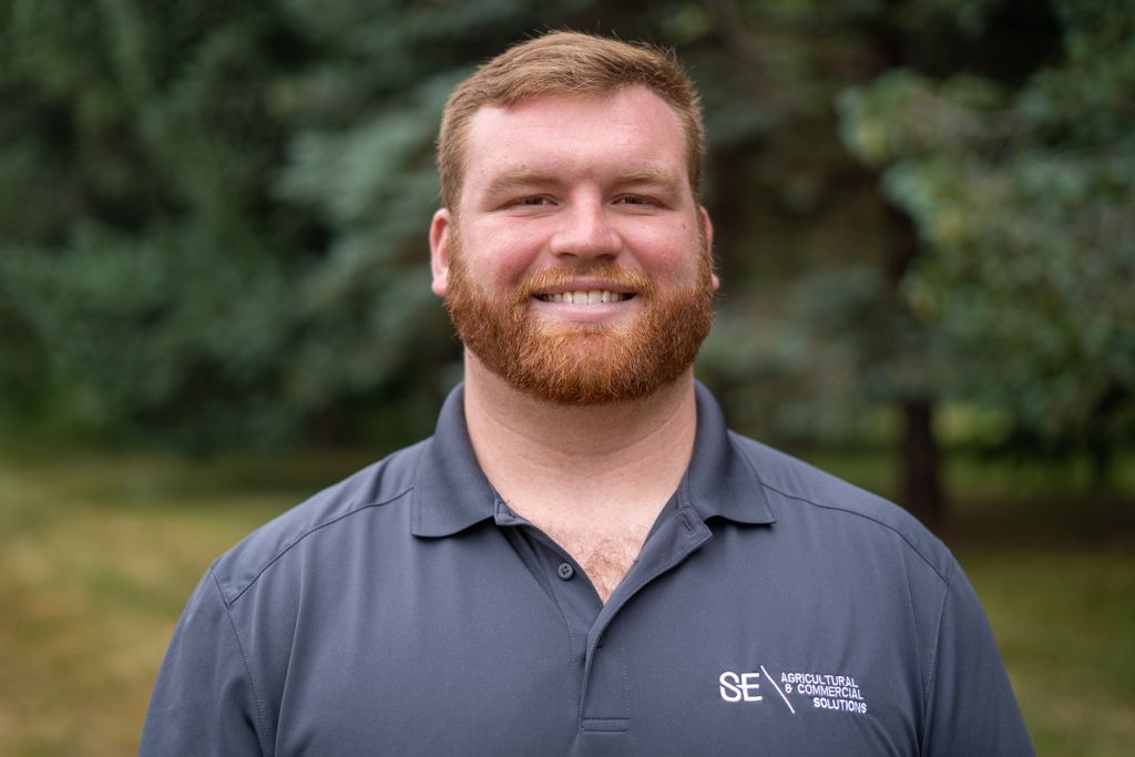 A person with reddish hair and a beard, wearing a gray polo shirt with a logo, smiling against a blurred green background.