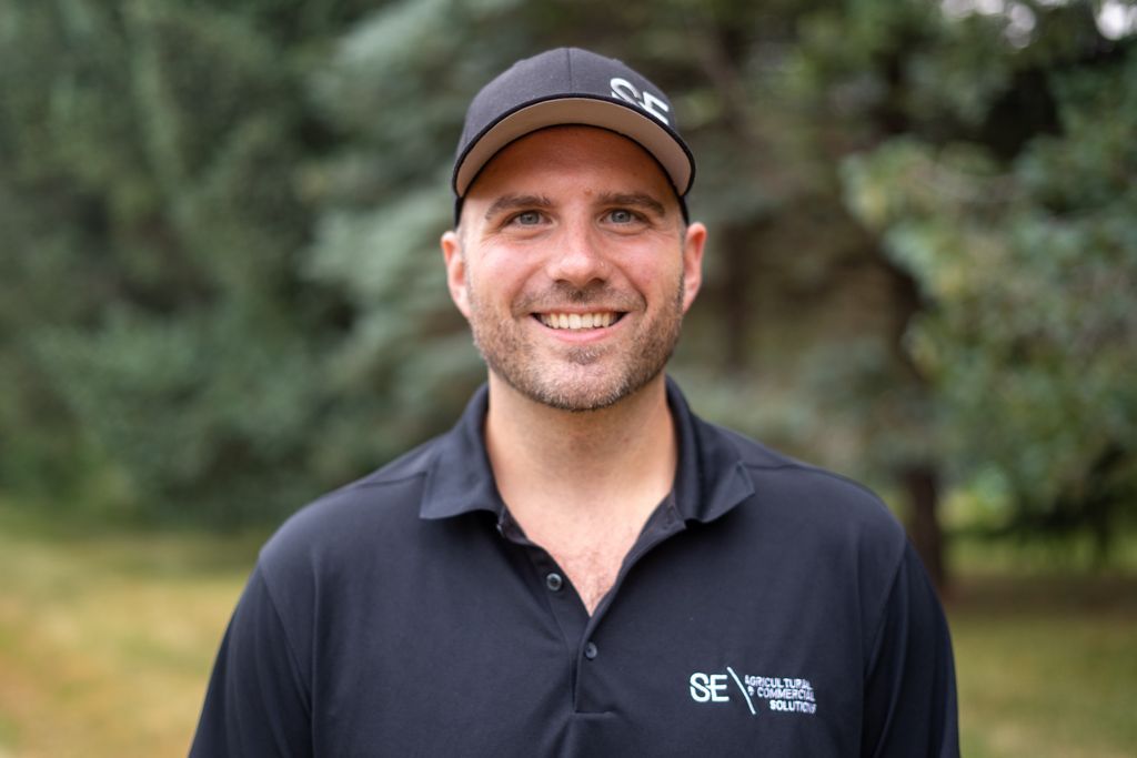 A smiling person wearing a black polo shirt and cap with a logo, standing in front of a blurred green, tree-filled background.