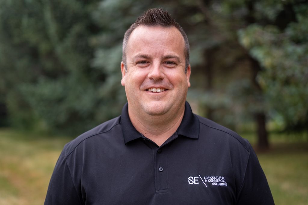 A man in a black polo shirt with a logo, smiling outdoors against a blurred background of green trees.