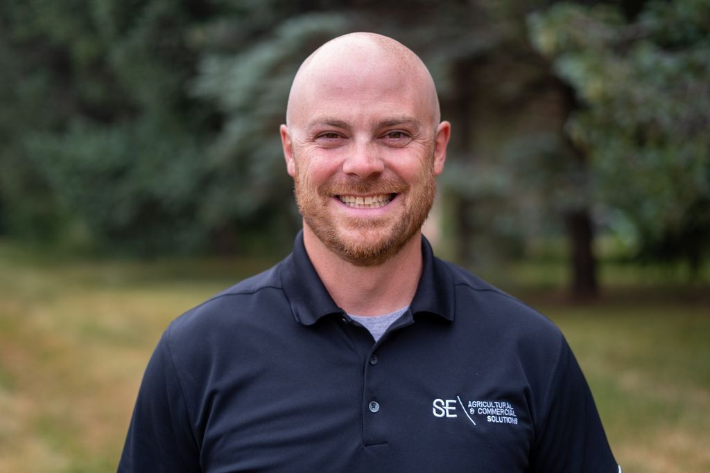 A smiling person with a shaved head and a beard, wearing a black polo shirt, standing in front of an out-of-focus park.