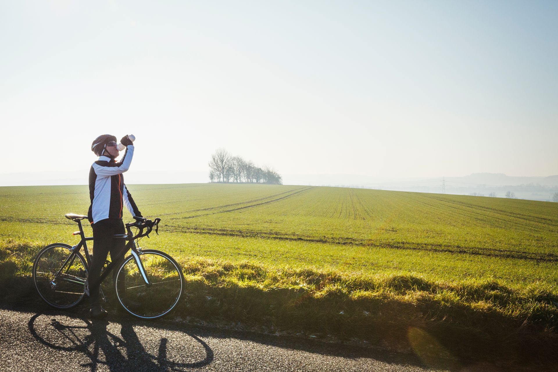 Ciclista bebiendo agua junto a su bicicleta en una carretera soleada, con vistas a un campo verde.