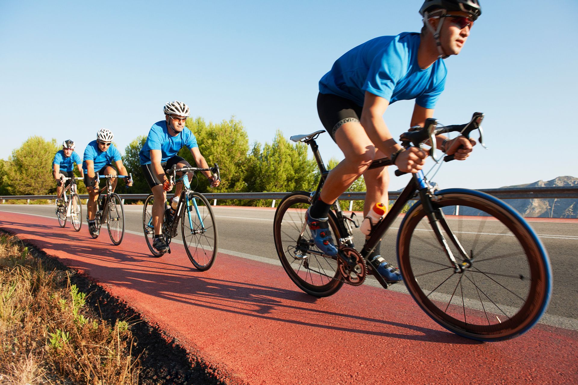 Ciclistas con camisetas azules montan en bicicleta en una pista roja al lado de una carretera, en un día soleado.