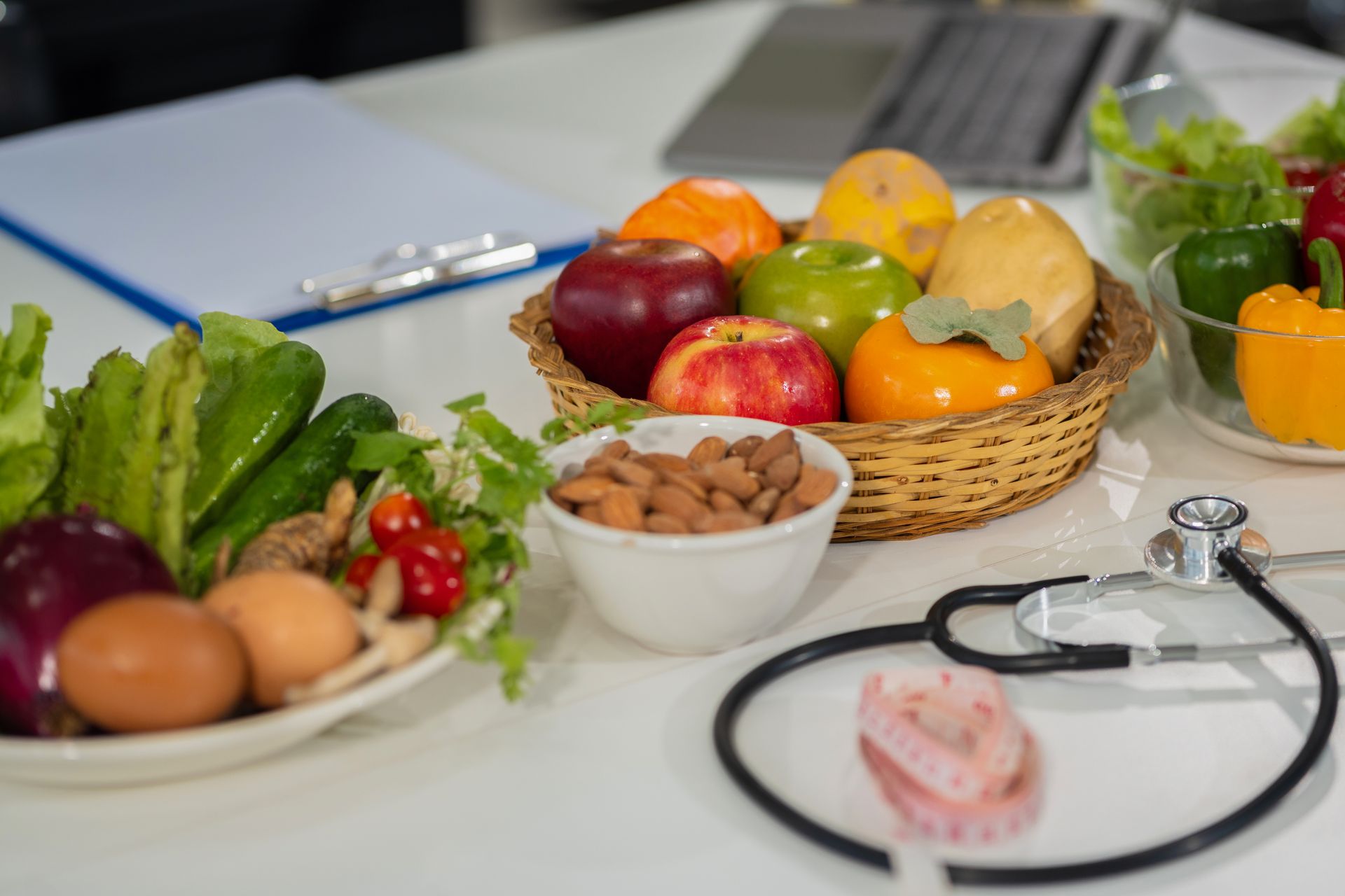 Disposición de alimentos saludables con frutas, verduras, frutos secos y un estetoscopio sobre una superficie blanca.