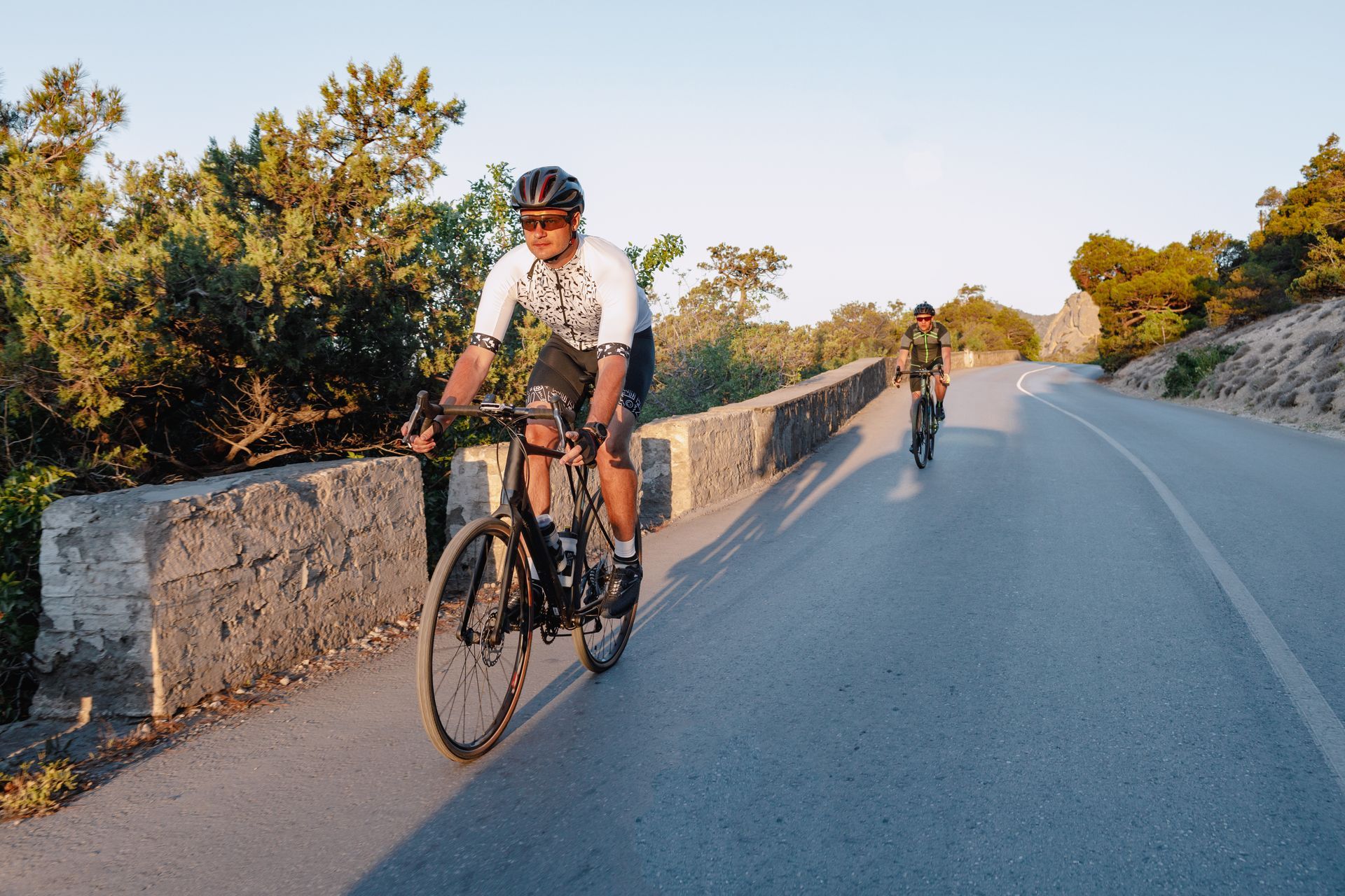 Ciclistas montando en bicicleta en una carretera pavimentada junto a un muro de piedra; árboles al fondo.