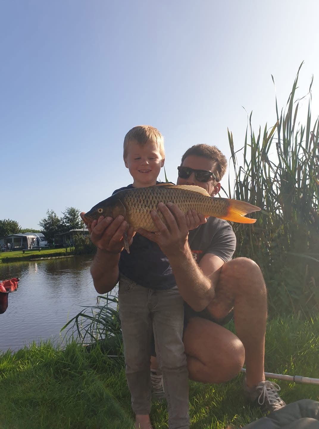 Mann und Kind halten einen großen Fisch an einem Teich; sonniger Tag.