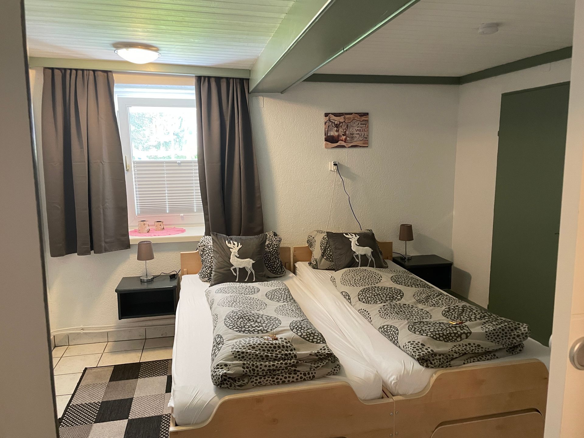 Ein Schlafzimmer mit zwei Betten, einem Fenster und dunklen Vorhängen. Graue und weiße Bettwäsche, schwarze Nachttische.