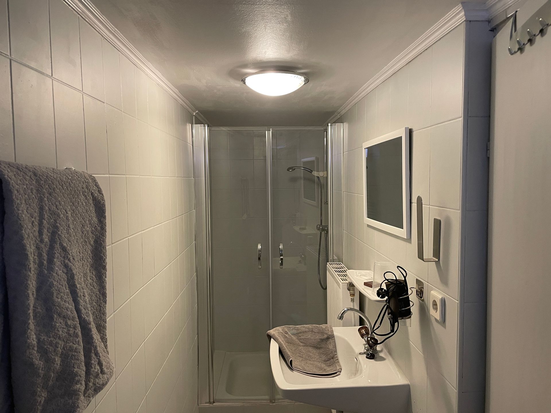 Kleines Badezimmer mit gefliesten Wänden, Dusche, Waschbecken, Spiegel und Handtuch.