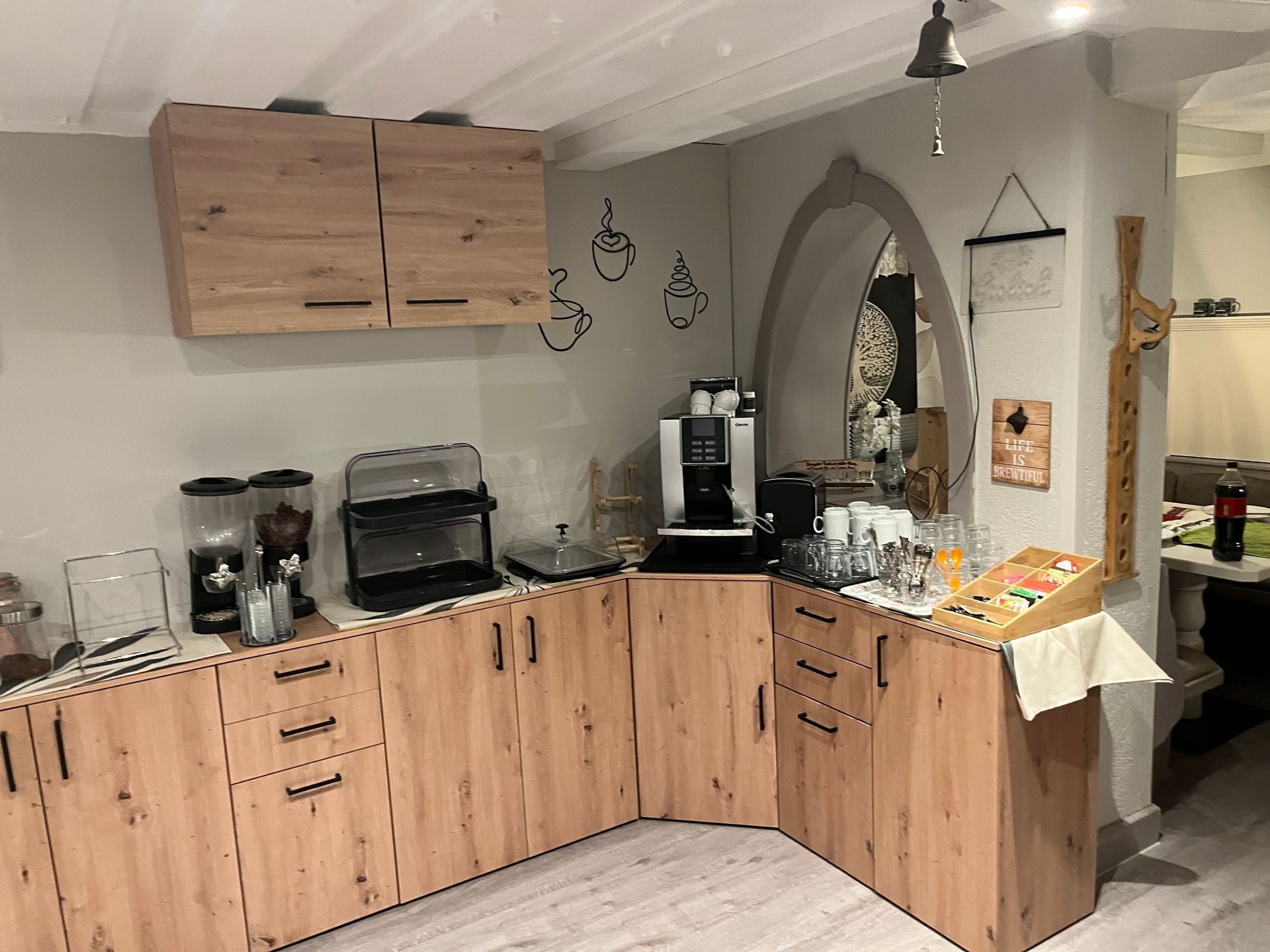 Küche mit Holzschränken, Elektrogeräten und einer Kaffeemaschine. Ein Torbogen führt in einen anderen Bereich.