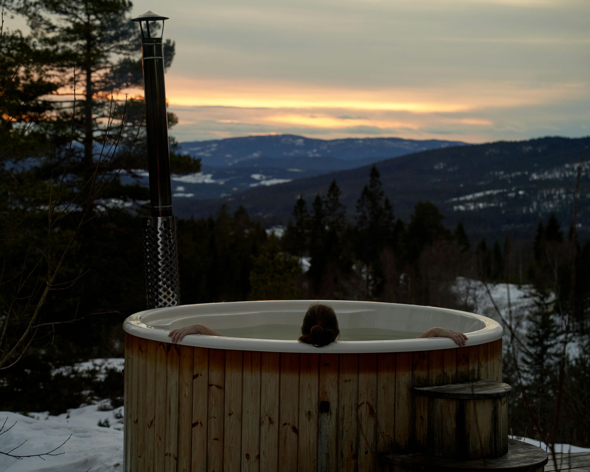  entspannt sich in einem hölzernen Whirlpool mit Blick auf einen verschneiten Wald und die Berge im Sonnenuntergang.