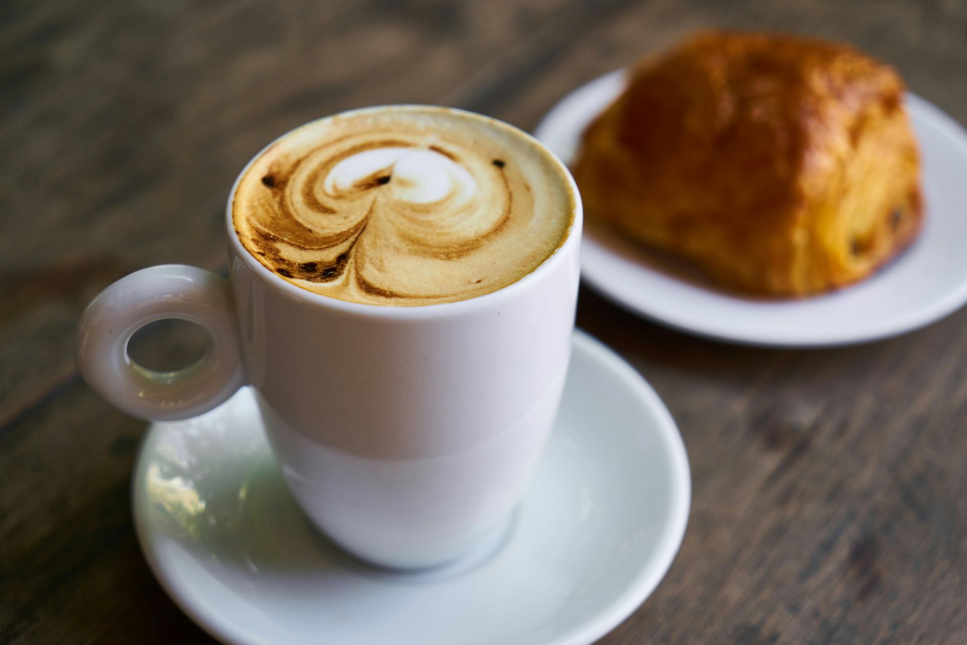 Cappuccino in weißer Tasse auf Untertasse; Gebäck auf weißem Teller, alles auf einem Holztisch.