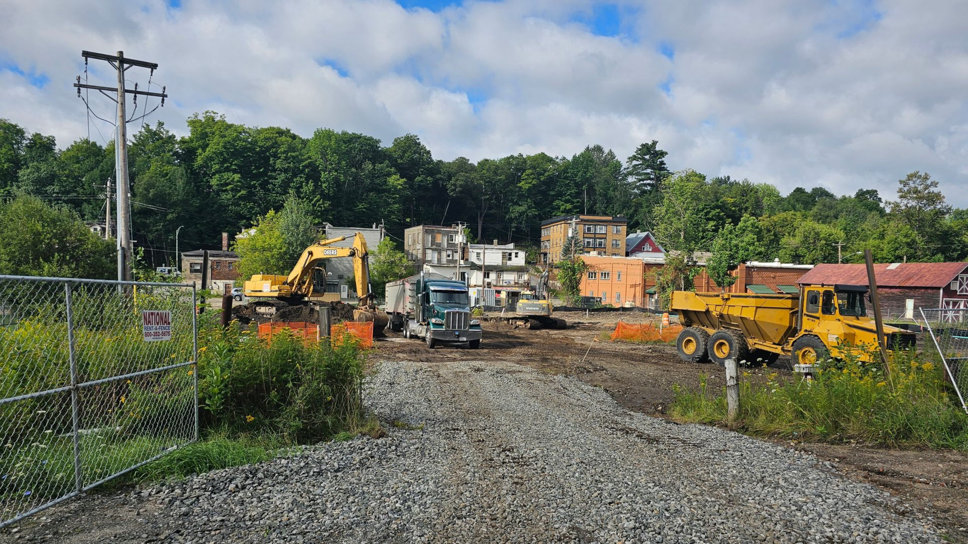 Construction site with heavy machinery, demolished buildings, and gravel road. Green trees and cloudy sky.