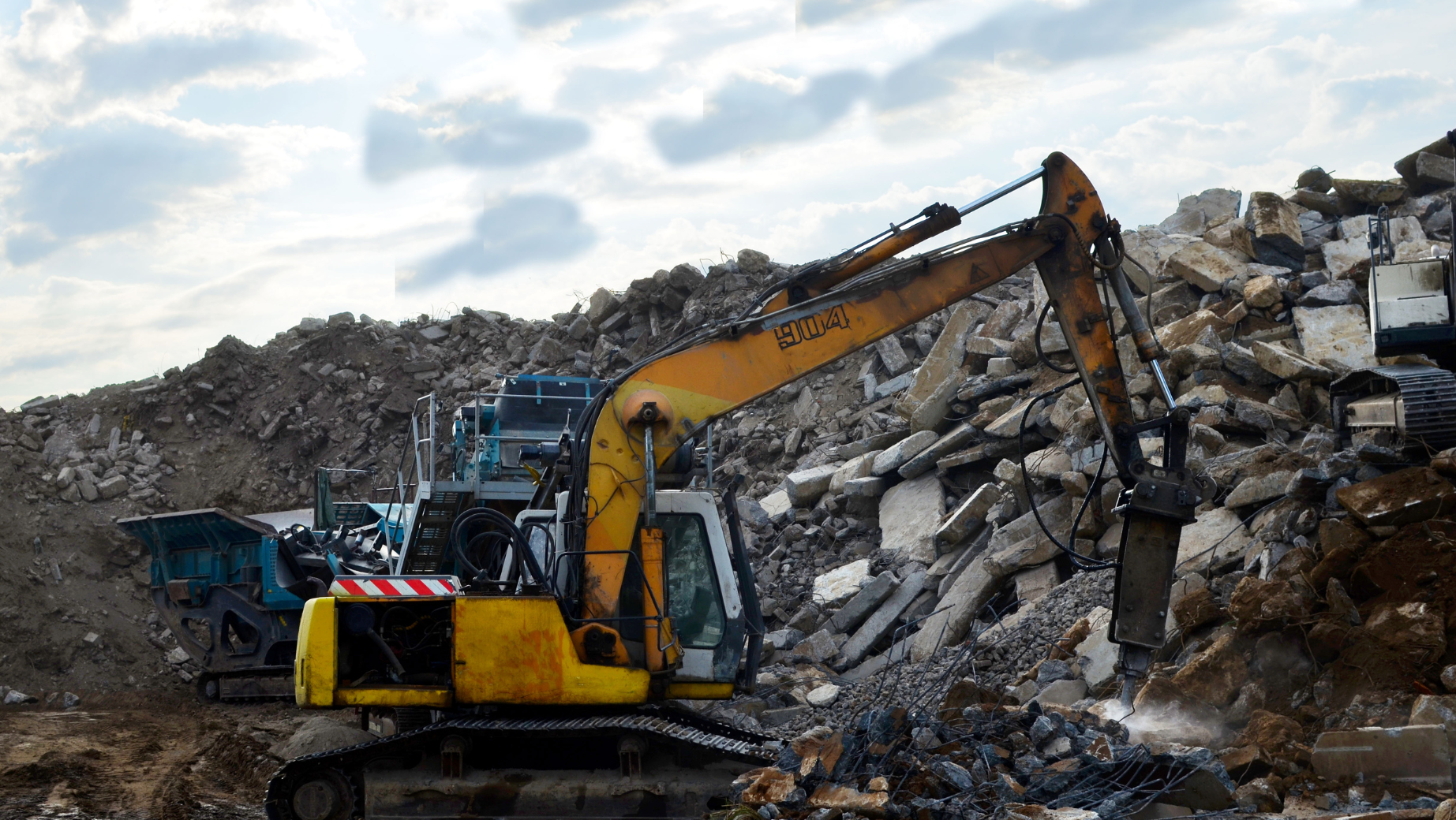 A yellow excavator is working on a pile of rubble and disposed building materials. 