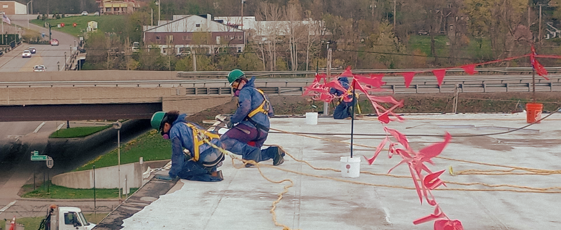 Workers wearing protective equipment and fall-arrest harnesses perform environmental remediation on a commercial rooftop.