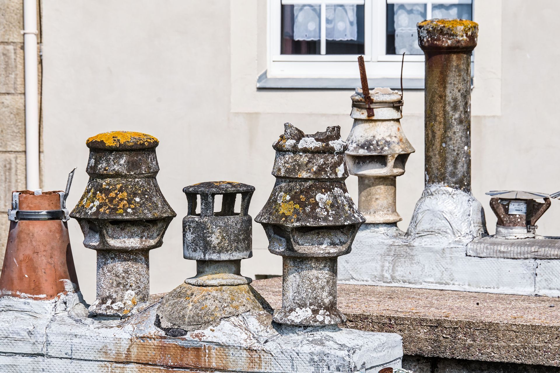 Weathered rooftop vent pipes and chimney stacks on an older building, showing corrosion, lichen growth, and patched roofing.