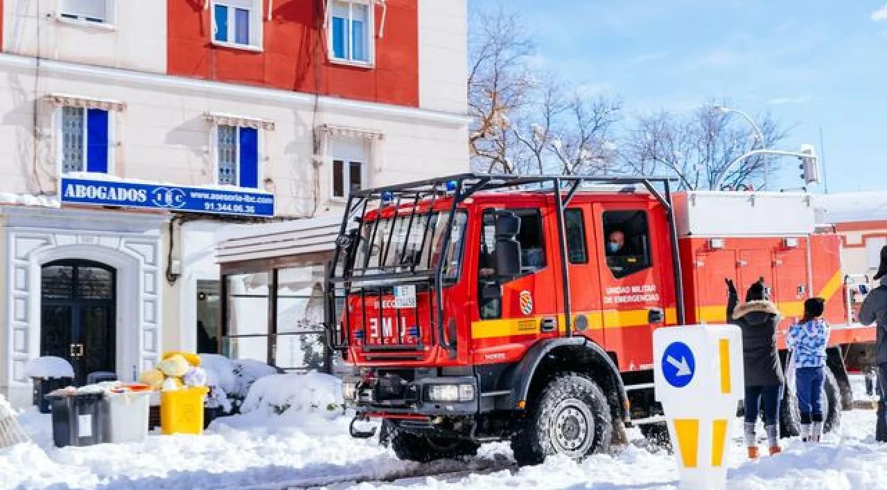 Red fire truck on a snowy street beside a building, with firefighters nearby.