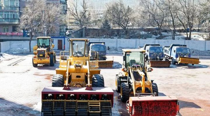 Snow-covered construction yard with yellow loaders and snowplows lined up in front of a fence.