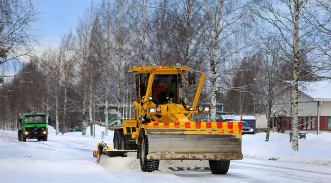 Snowplow clearing a snowy street, with a green vehicle in the distance and bare trees lining the road.