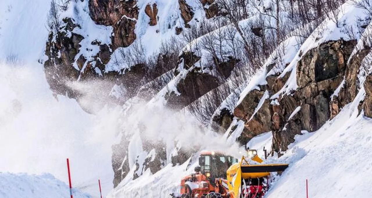 Snowplow clearing a snowy mountain road beside rock cliffs, with white snow and equipment in view