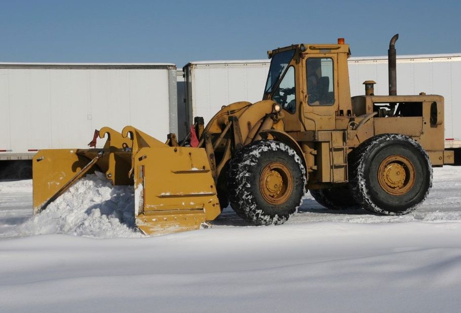 Yellow snowplow clearing snow on a snowy lot beside white trailers