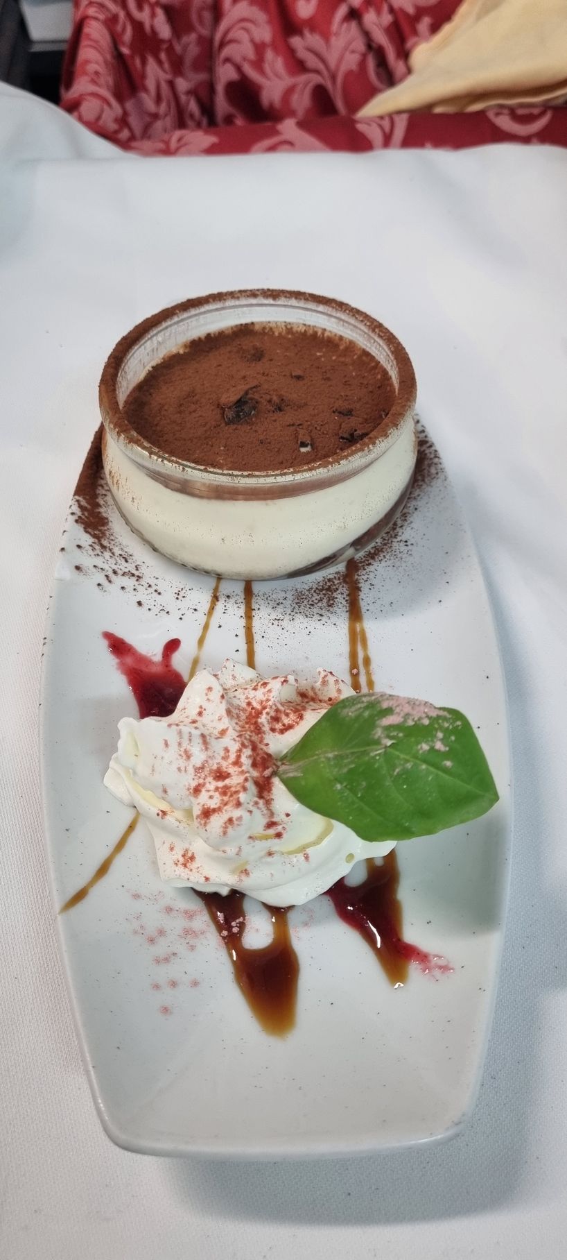 Dessert tiramisù con panna montata, salsa rossa e una foglia su un piatto bianco.