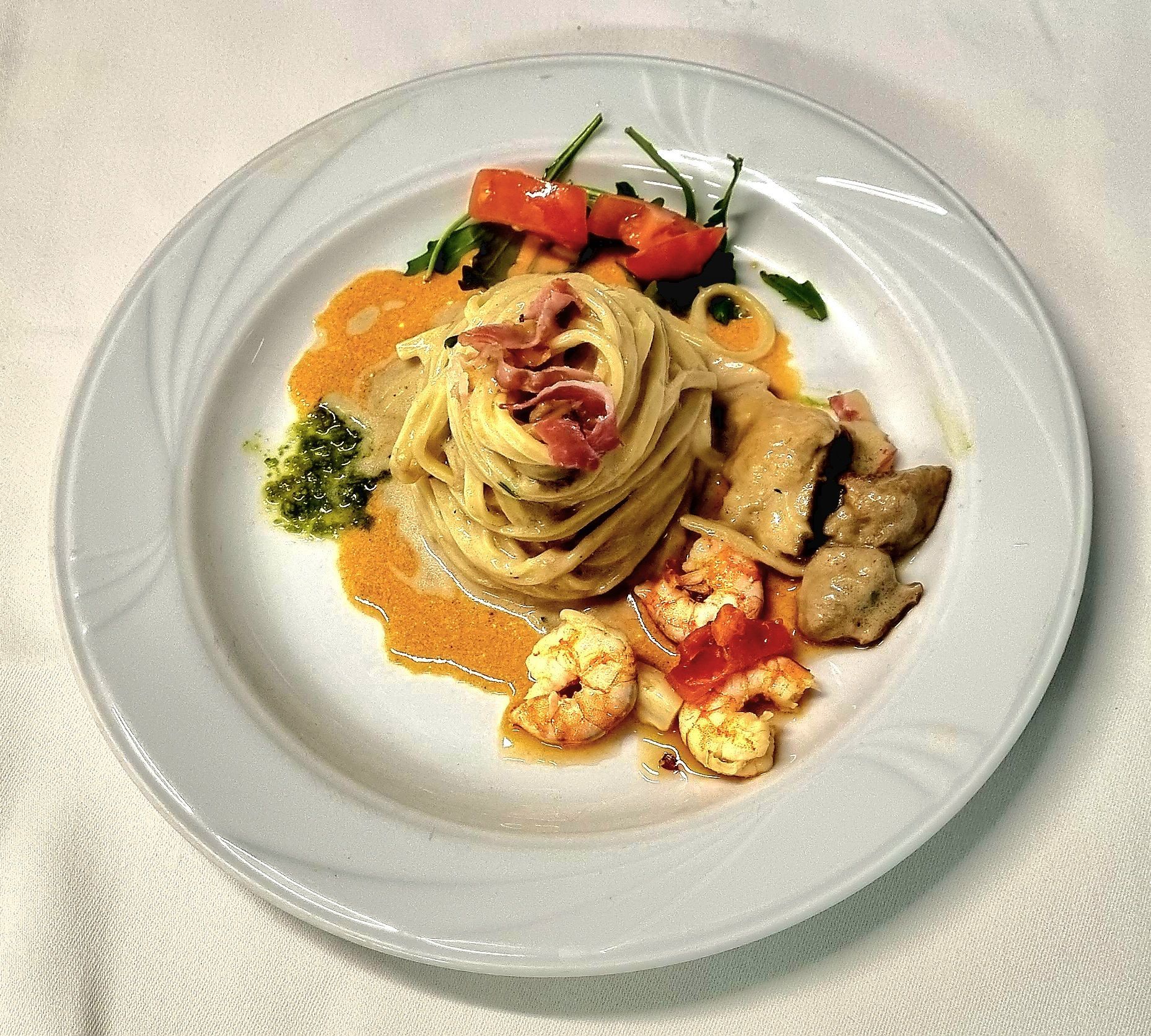 Piatto di pasta con gamberi, funghi e prosciutto su un piatto bianco, guarnito con verdure e pomodori.