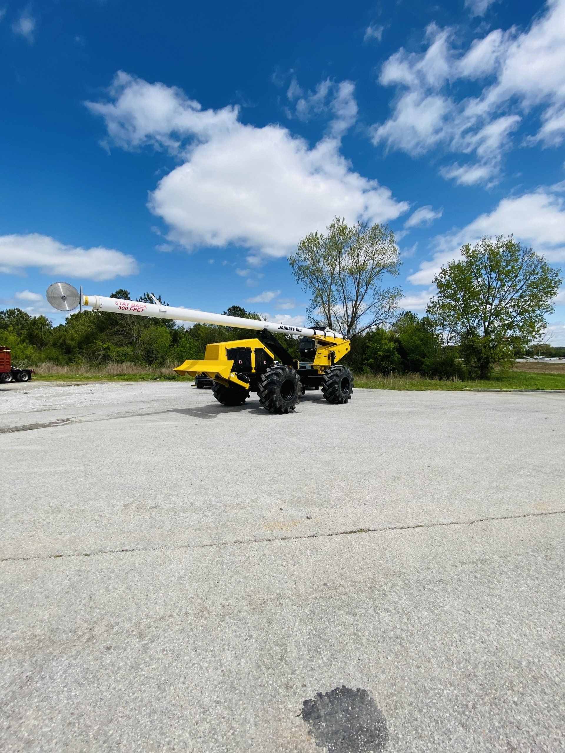 Yellow forklift on a paved lot under a blue sky with clouds, trees in the background