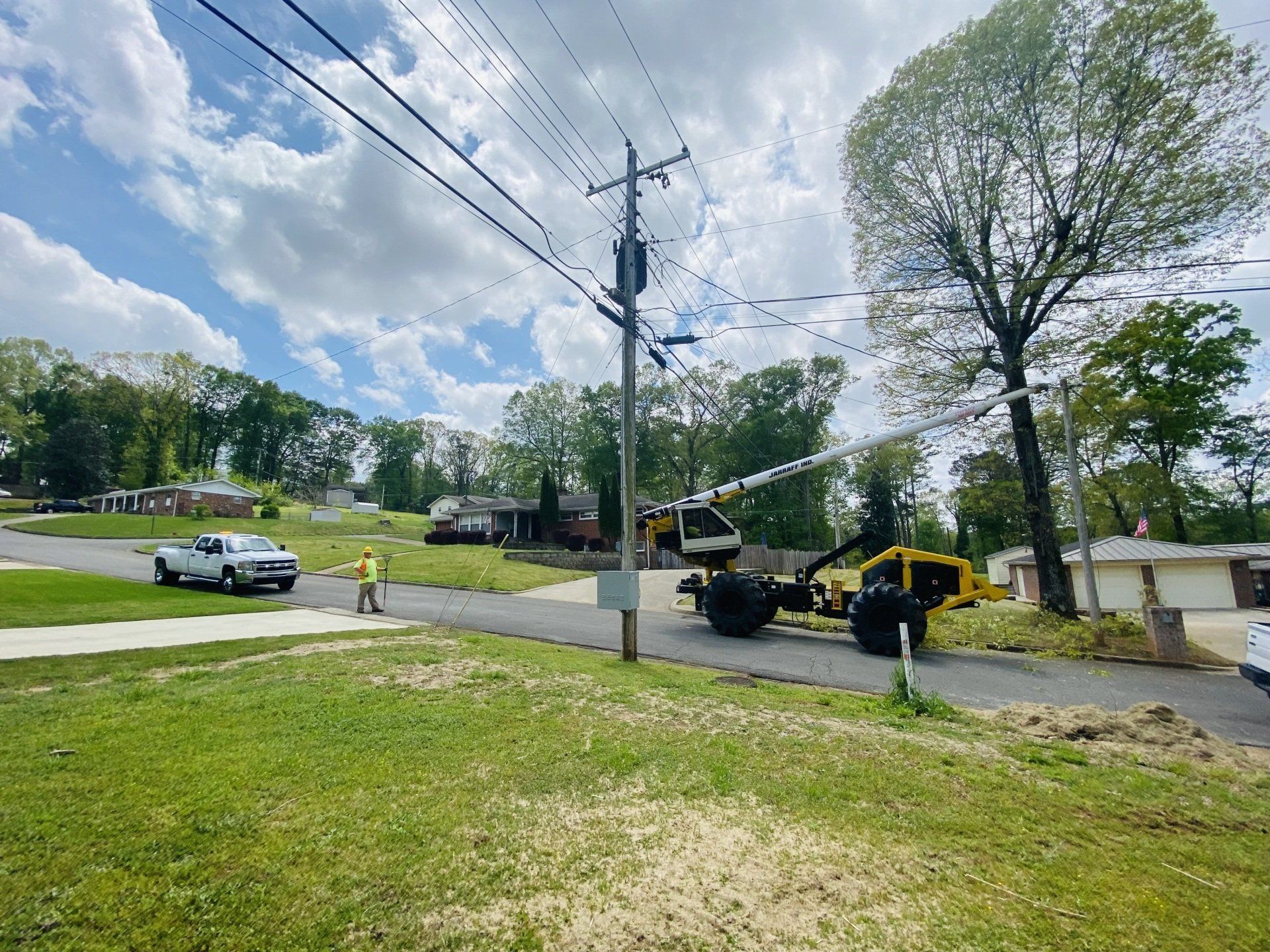 Construction equipment and workers beside a road in a grassy suburban neighborhood under a cloudy sky