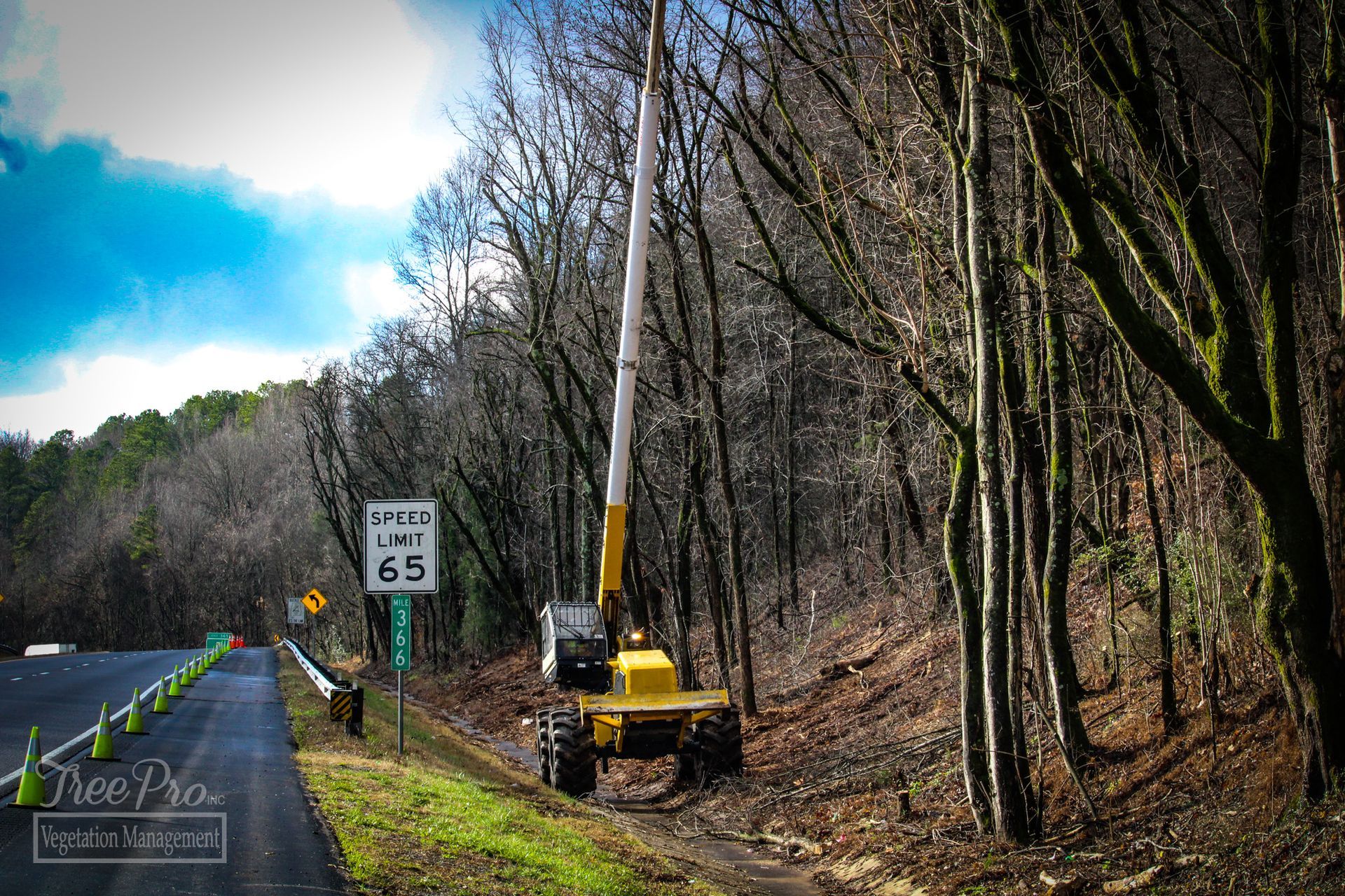 Yellow utility truck trimming trees beside a roadside speed limit 65 sign on a wooded hillside