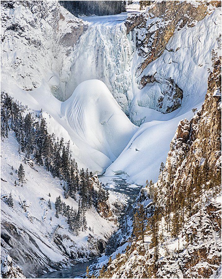 A waterfall is surrounded by snow covered mountains and trees.