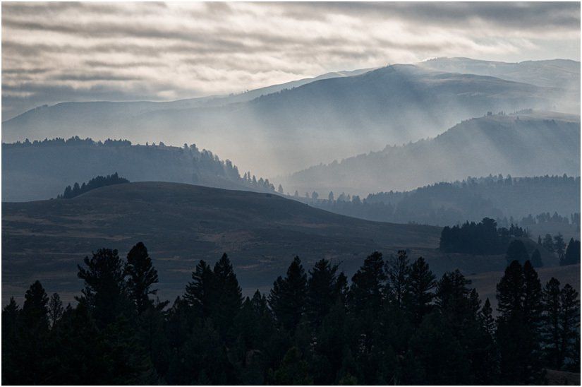 The sun is shining through the clouds over the mountains and trees.