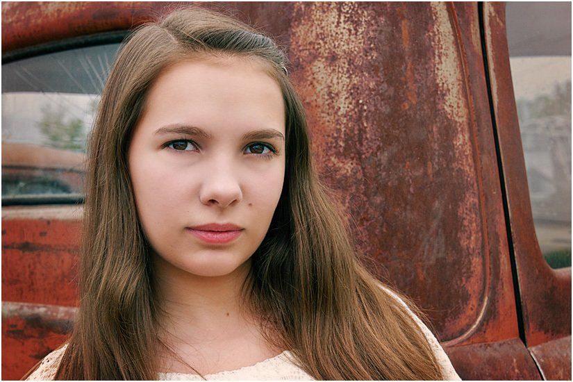 A young girl is standing in front of a rusty truck.