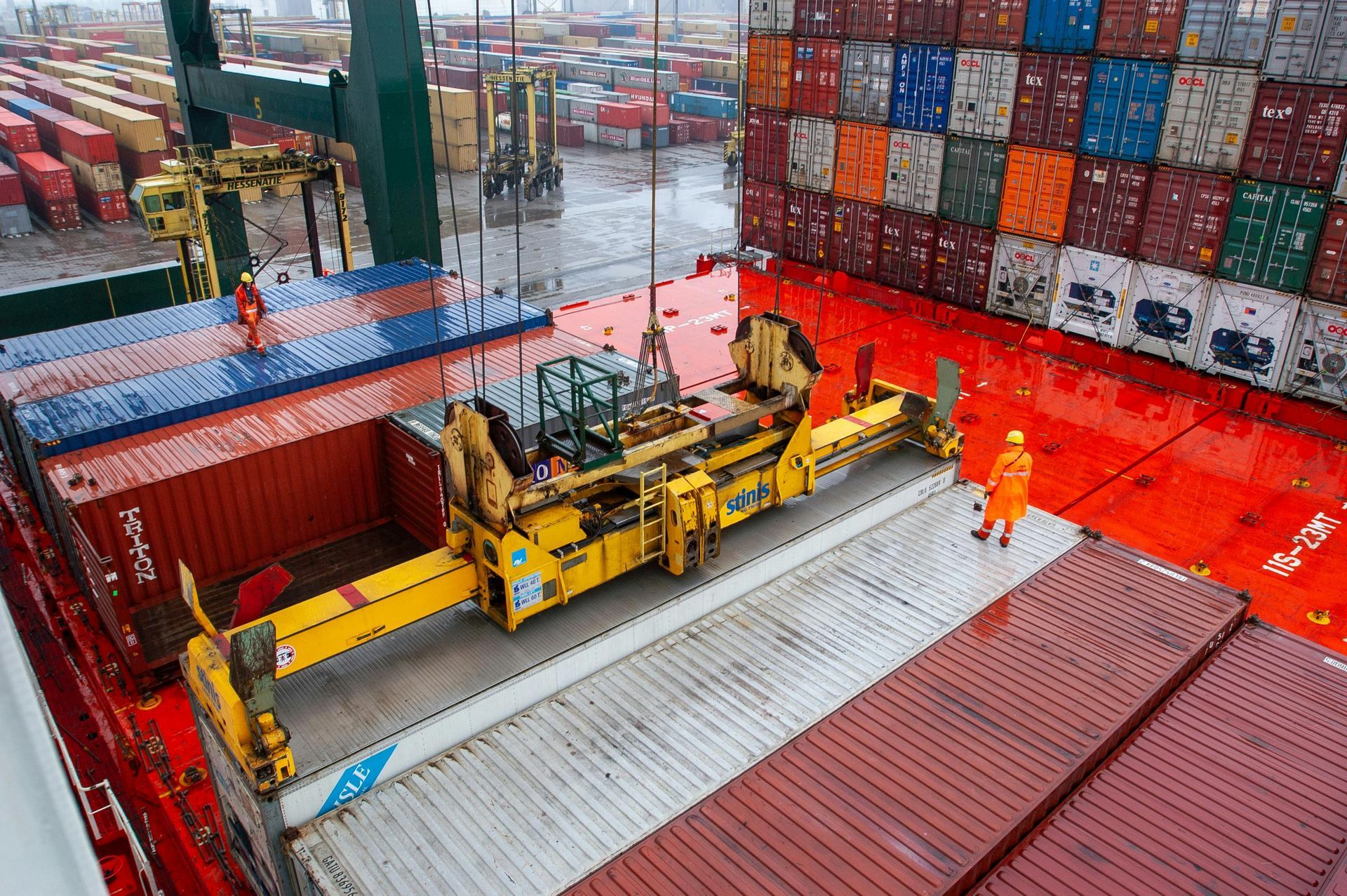 Workers in safety gear operate a crane lifting a shipping container onto a cargo ship deck surrounded by stacked containers at a busy port
