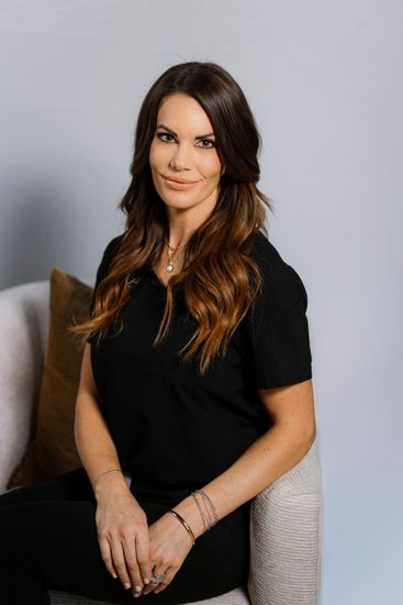 Woman seated on a chair, wearing a black dress, with long brown hair and a neutral studio backdrop