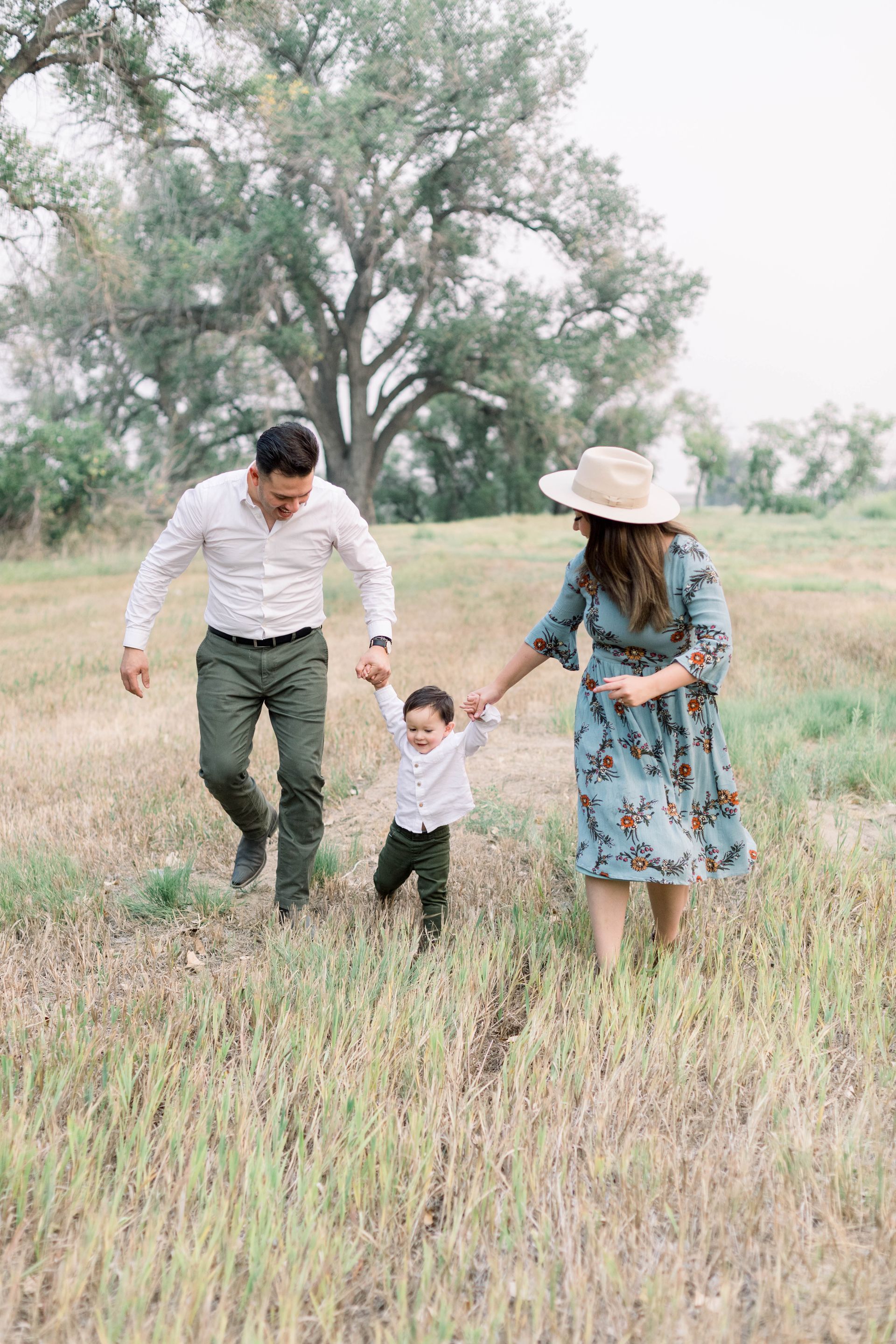 Family walking in a field holding hands. Man in white shirt and woman in floral dress, leading a child.