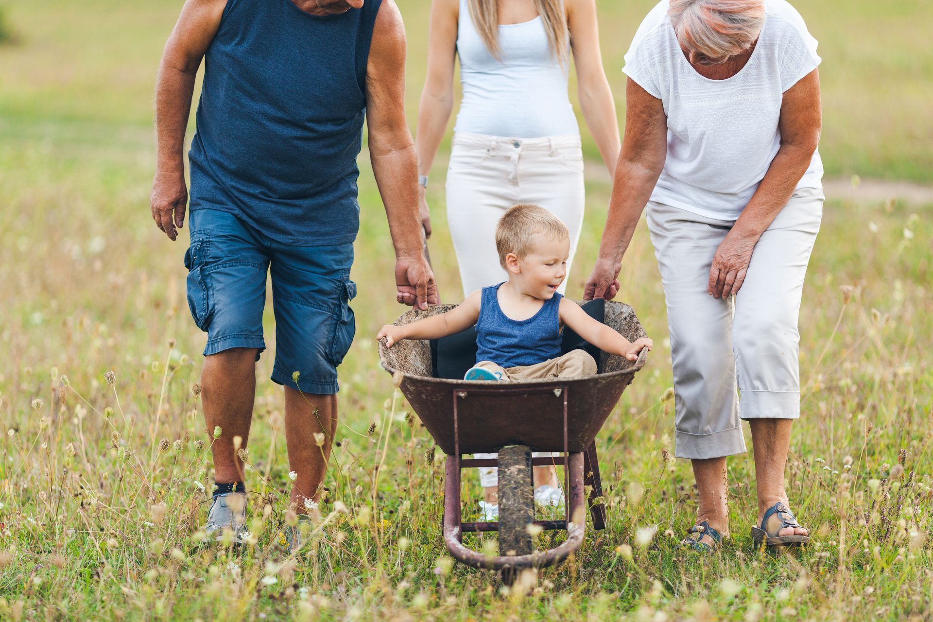 A young child sits in a wheelbarrow, pushed through a grassy field by two adults.