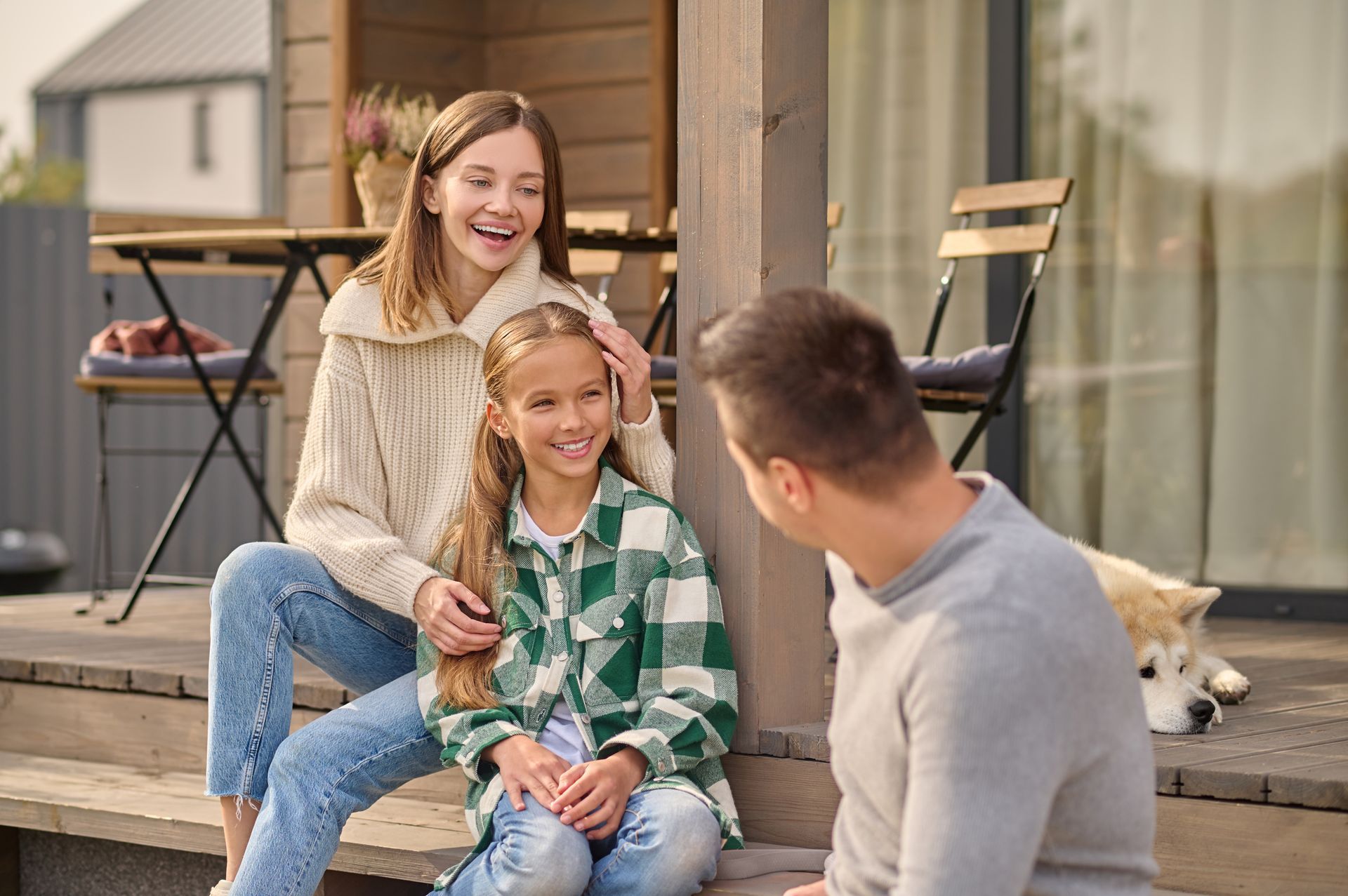 Family on porch, smiling. Woman and girl sit on steps; man stands near. Dog rests nearby.