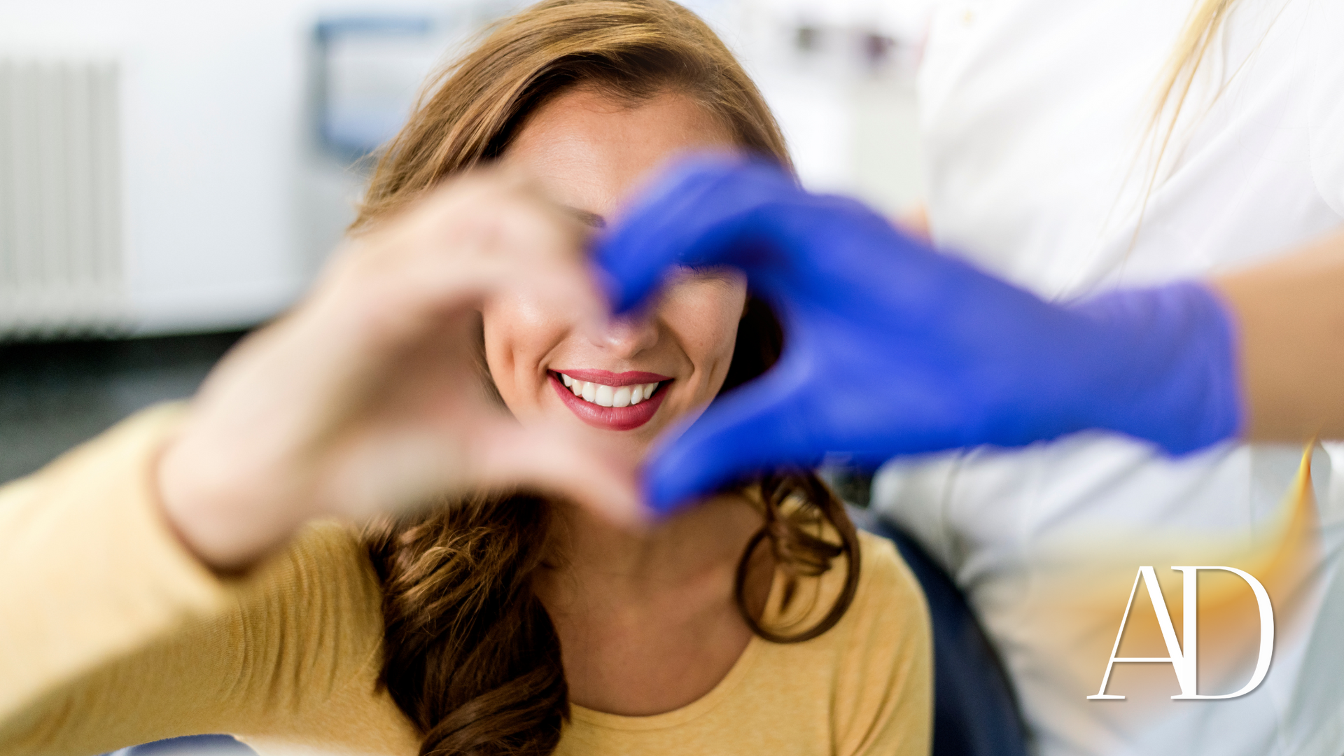 Woman smiling as gloved hands frame a heart shape over her face in a bright clinic setting